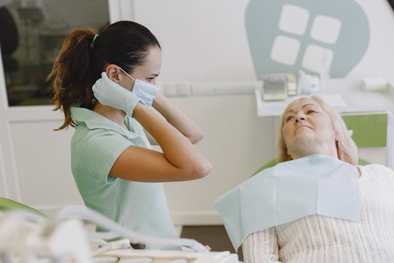 A dentist wearing a mask prepares to attend to an elderly patient lying down.