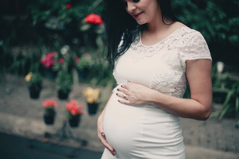 Pregnant woman in white lace dress, posing outdoors with flowers, exuding happiness.